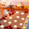 A tray of floating candles in a glass pan surrounded by red rose petals and wood accents.
