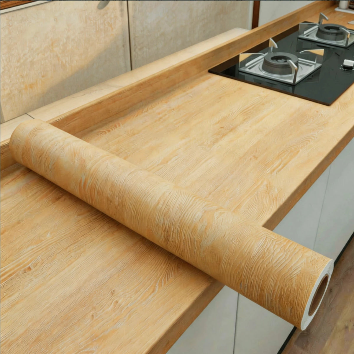A roll of self-adhesive wood grain wallpaper used as a cutting mat on a light wooden kitchen countertop.