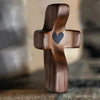 A dark wooden comfort prayer cross with a small heart carved into the center, resting on a textured surface.