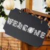 A black sign with white floral alphabet stencil letters spelling 'WELCOME' is displayed on a wooden tray.