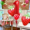 A red and green strawberry foil balloon decoration set for a first birthday party.