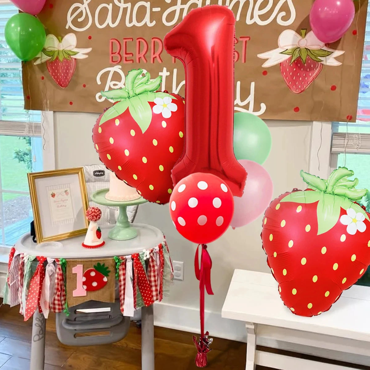A red and green strawberry foil balloon decoration set for a first birthday party.