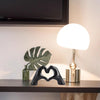 A black heart-shaped resin hand sculpture sits on a wooden shelf next to a modern lamp.
