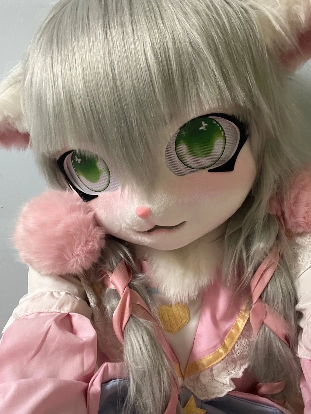 A light gray furry cosplay head with large green eyes and pink accents.
