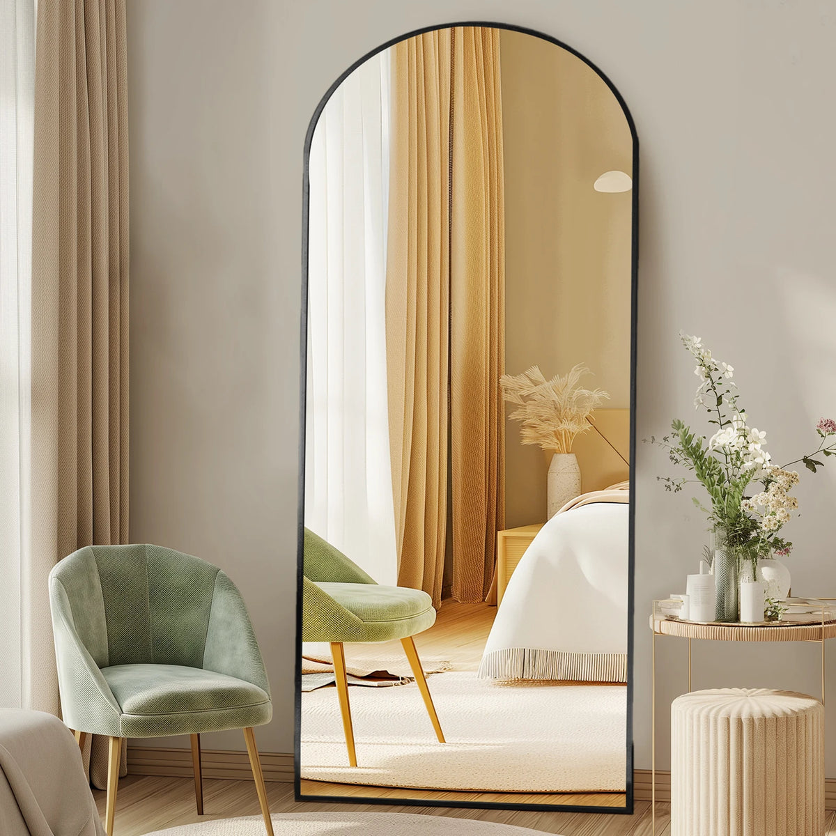 A full length arched wall mirror reflects a bedroom with beige curtains and a green armchair.