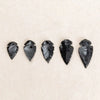Five polished black obsidian arrow pendants arranged horizontally on a light beige background.