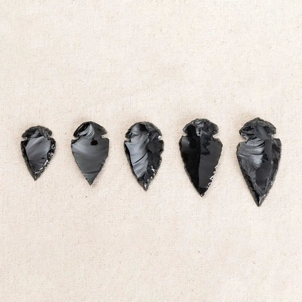 Five polished black obsidian arrow pendants arranged horizontally on a light beige background.