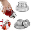 A hand holds a glass jar with an orange liquid, next to a stainless steel shaker lid and a glass filled with liquid and herbs