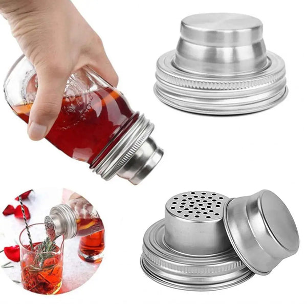 A hand holds a glass jar with an orange liquid, next to a stainless steel shaker lid and a glass filled with liquid and herbs