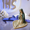 A statue of the Virgin Mary with a green mantle and rosary beads rests on a light-colored tray.