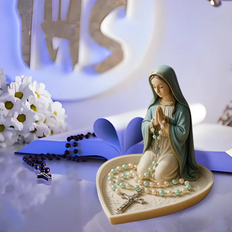 A statue of the Virgin Mary with a green mantle and rosary beads rests on a light-colored tray.