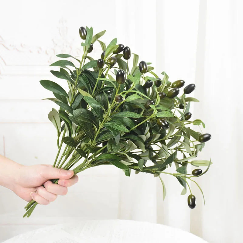 A hand holds an artificial olive leaf branch with dark green leaves and olives for home or wedding decor.