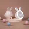 A pink mini LED cartoon alarm clock night light with rabbit ears sitting on a light wooden stand.