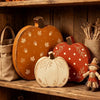 A set of three wooden pumpkin figurines in orange, red, and white are displayed on a wooden shelf.