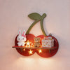 A wooden shelf shaped like a cherry with a green leaf, holding a white rabbit and a teddy bear.