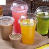 A collection of colorful fruit juices and smoothies in clear plastic cups with dome lids displayed on a burlap surface.