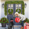 A woman in a beige coat is standing in front of a dark blue door decorated with a green cedar garland and wreaths.