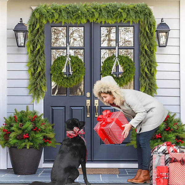 A woman in a beige coat is standing in front of a dark blue door decorated with a green cedar garland and wreaths.