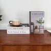A stack of books featuring the title Create at Home with Old & New and Pacific Natural at Home displayed on a wooden surface.