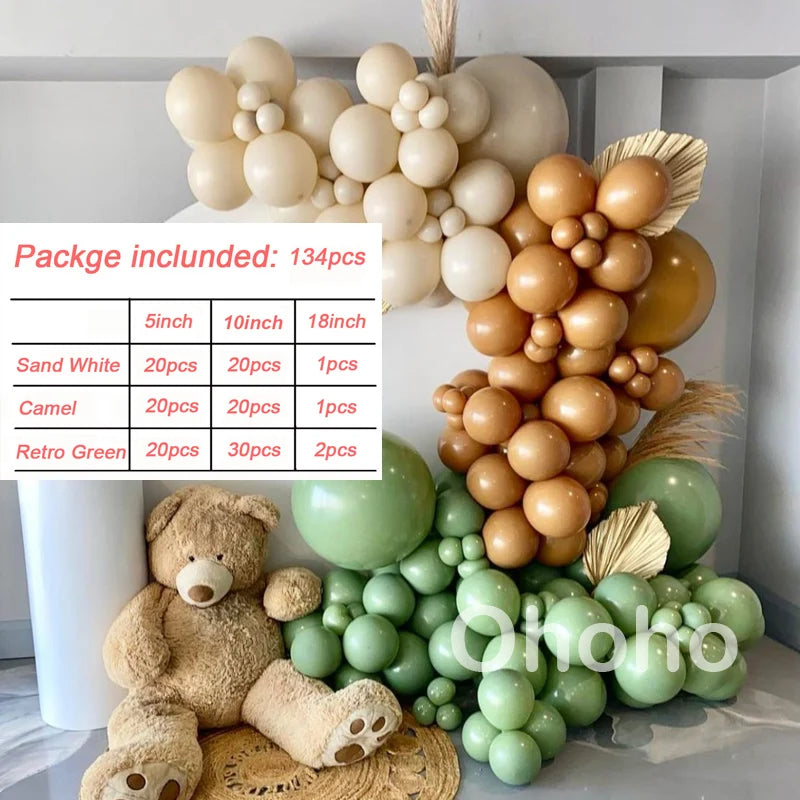 A balloon garland kit featuring beige and gold balloons arranged on a white shelf with a teddy bear and green balloons below.