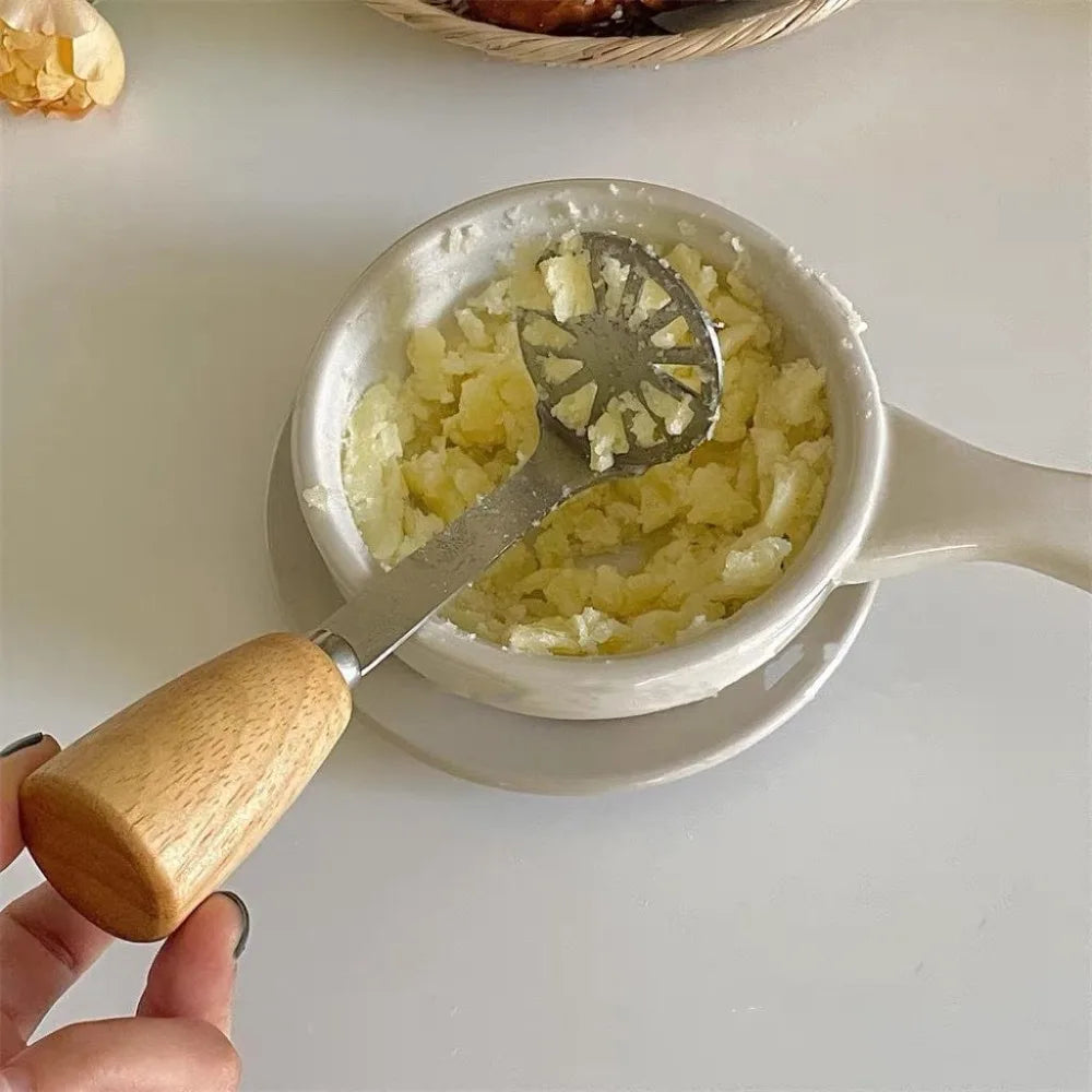 A hand holds a manual wooden potato and pumpkin masher with a wooden handle and metal blade, positioned above a bowl of mashe