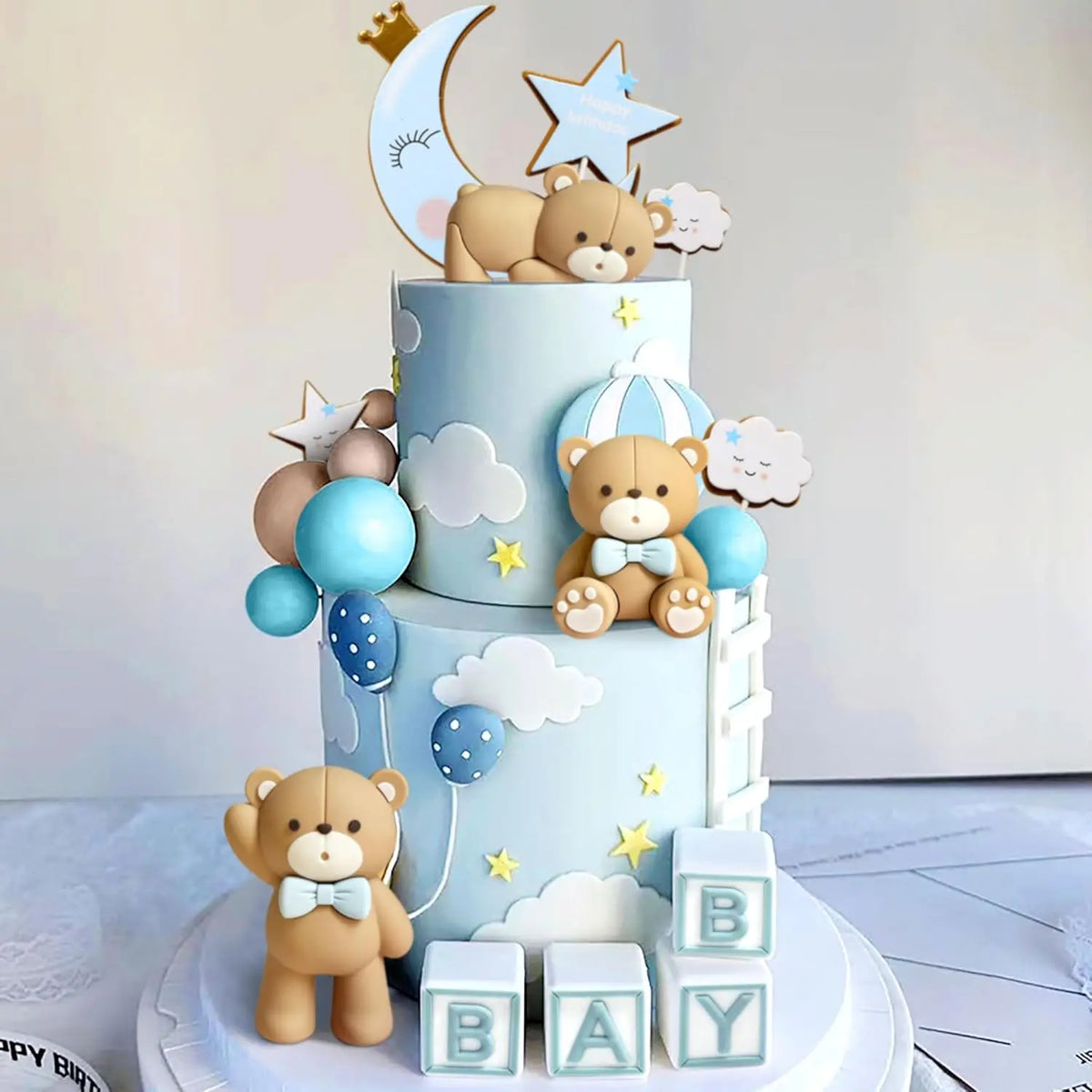 A tiered baby shower cake featuring a bear topper, stars, clouds, and blue balloons on a white background.