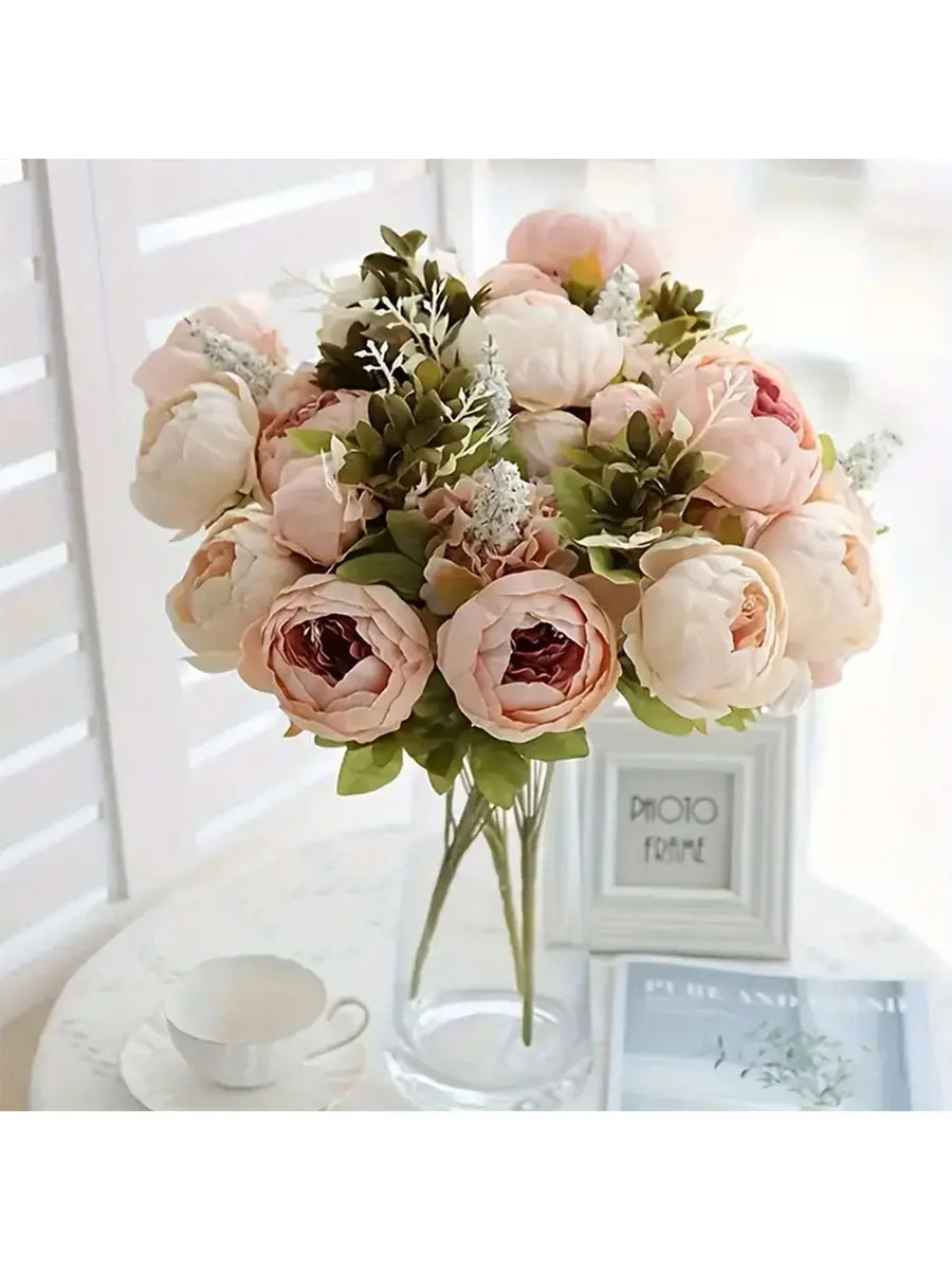 A bouquet of pink and cream peony artificial flowers with green foliage displayed in a glass vase.