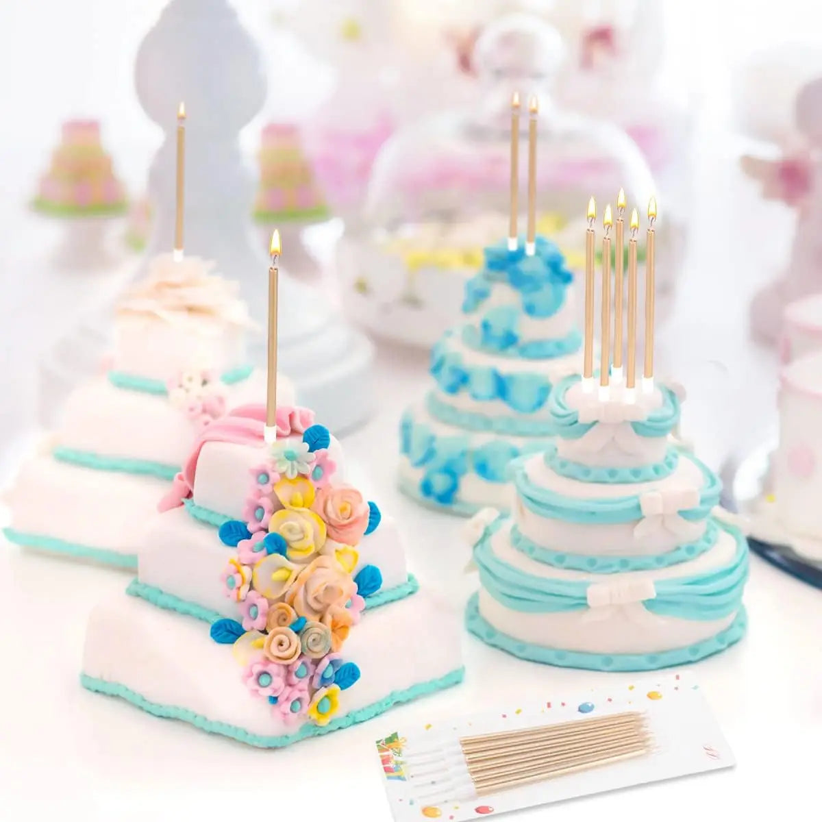 A display of metallic birthday cake candles with holders, featuring tiered cakes decorated in light blue and pink hues.