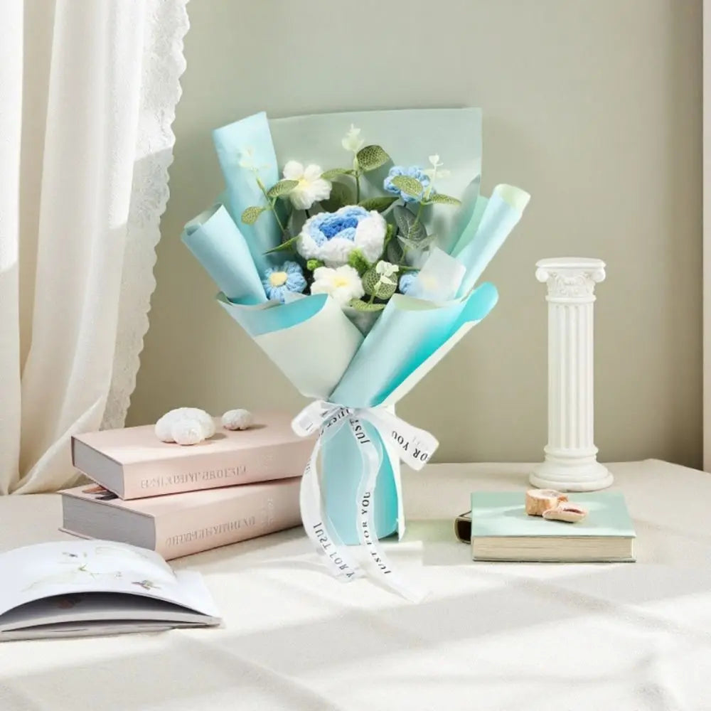 A handmade crochet rose flower bouquet wrapped in light blue paper and presented with stacked books.