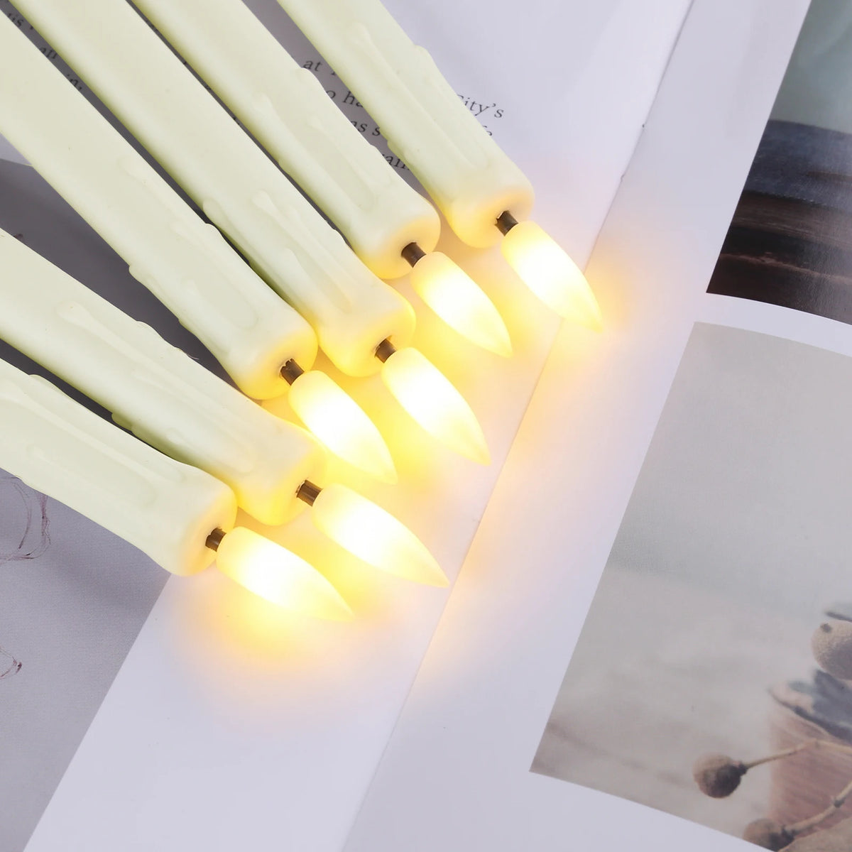A close-up of six white flameless taper candles with glowing yellow tips arranged diagonally on a white surface.