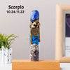 A blue crystal bottle shaped like a scorpion with raw healing stones, displayed on a wooden surface.
