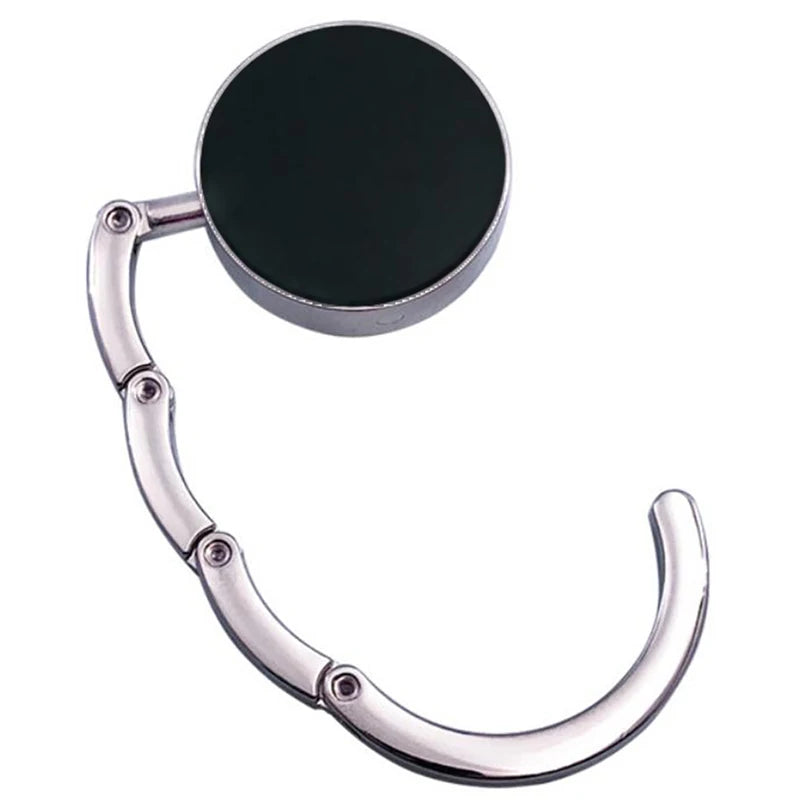 A silver foldable handbag hook with a round black plate, designed for hanging bags on tables and desks.