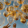 A close-up view of numerous gold and iridescent confetti balloons floating against a clear blue sky.