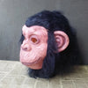 A realistic latex monkey mask with pinkish skin and black fur, displayed against a gray background.