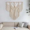 A beige macrame wall hanging tapestry with fringe and tassels is displayed on a white wall above a light-colored sofa.