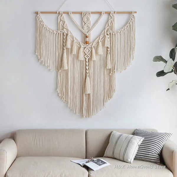 A beige macrame wall hanging tapestry with fringe and tassels is displayed on a white wall above a light-colored sofa.