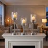 Three silver metal pillar candle holders with lit candles displayed on a white mantelpiece.
