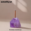 A purple amethyst crystal sits on a surface with a wooden stick and a small metal tip, used as an aromatherapy holder.