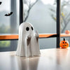 A white ghost figurine with an expressive hand gesture sits on a wooden desk near a pumpkin.