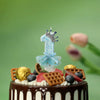 A birthday cake decorated with a blue crown candle and light blue fabric skirt on a green background.