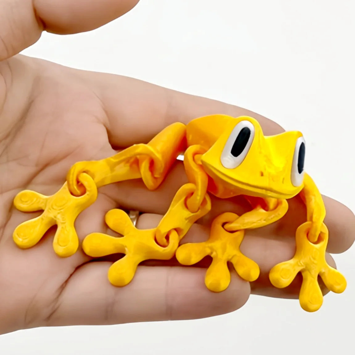 A hand holds a yellow, realistic movable 3D printed frog fridge magnet.