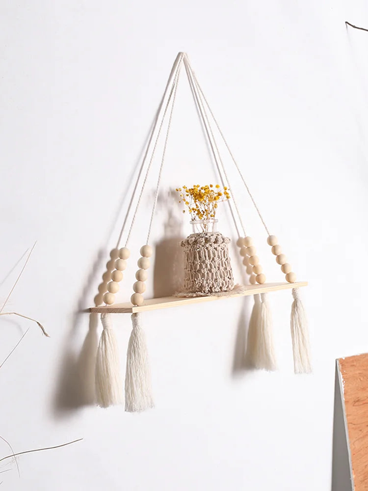 A rustic wooden wall shelf with macrame hangers displaying dried yellow flowers and white tassels.