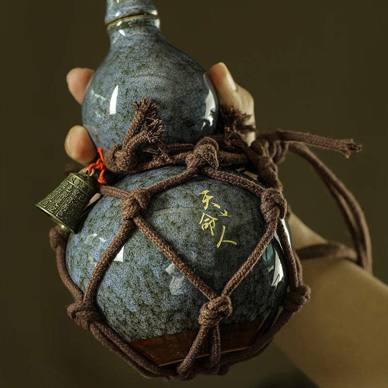 A hand holds a dark blue ceramic wine gourd with a woven cord and a small brass tag.
