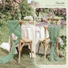 A rustic outdoor wedding setting featuring sage green gauze chair sashes and a white table runner.