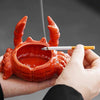A hand holds a red ceramic crab ashtray with a lid, showing the interior and an lit cigarette resting on the rim.