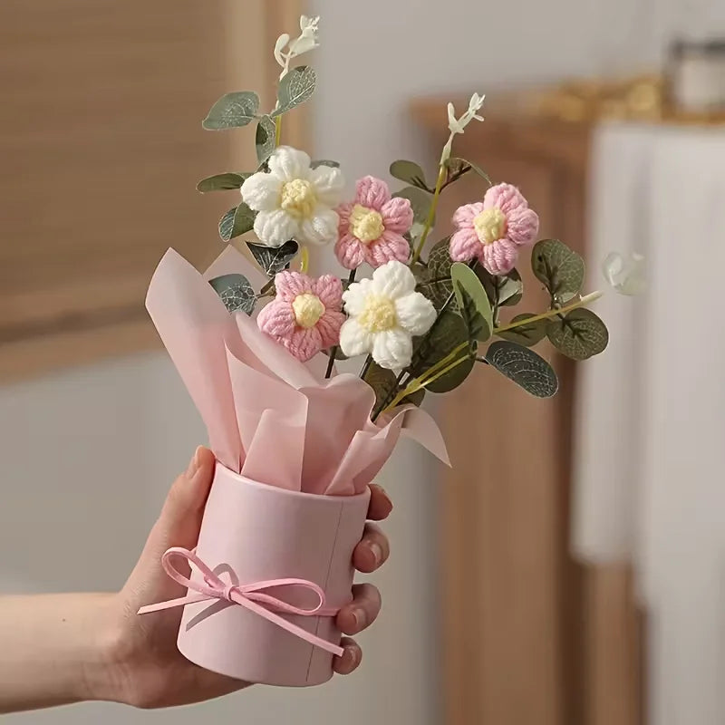 A hand holds a pink paper bouquet featuring white and pink flowers with green leaves.