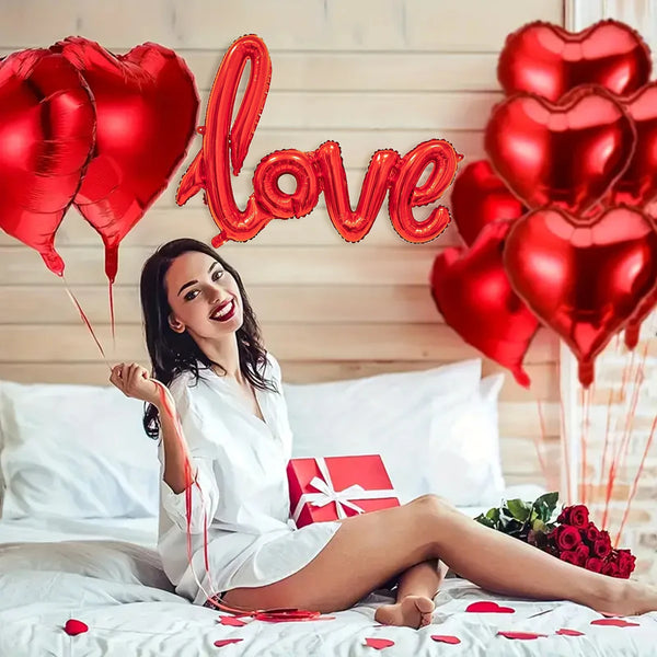 A woman smiling while lying on a bed surrounded by red heart-shaped balloons and scattered red petals.