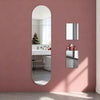 A vertical oval mirror reflects a white shelf with decorative items mounted on a pink wall.