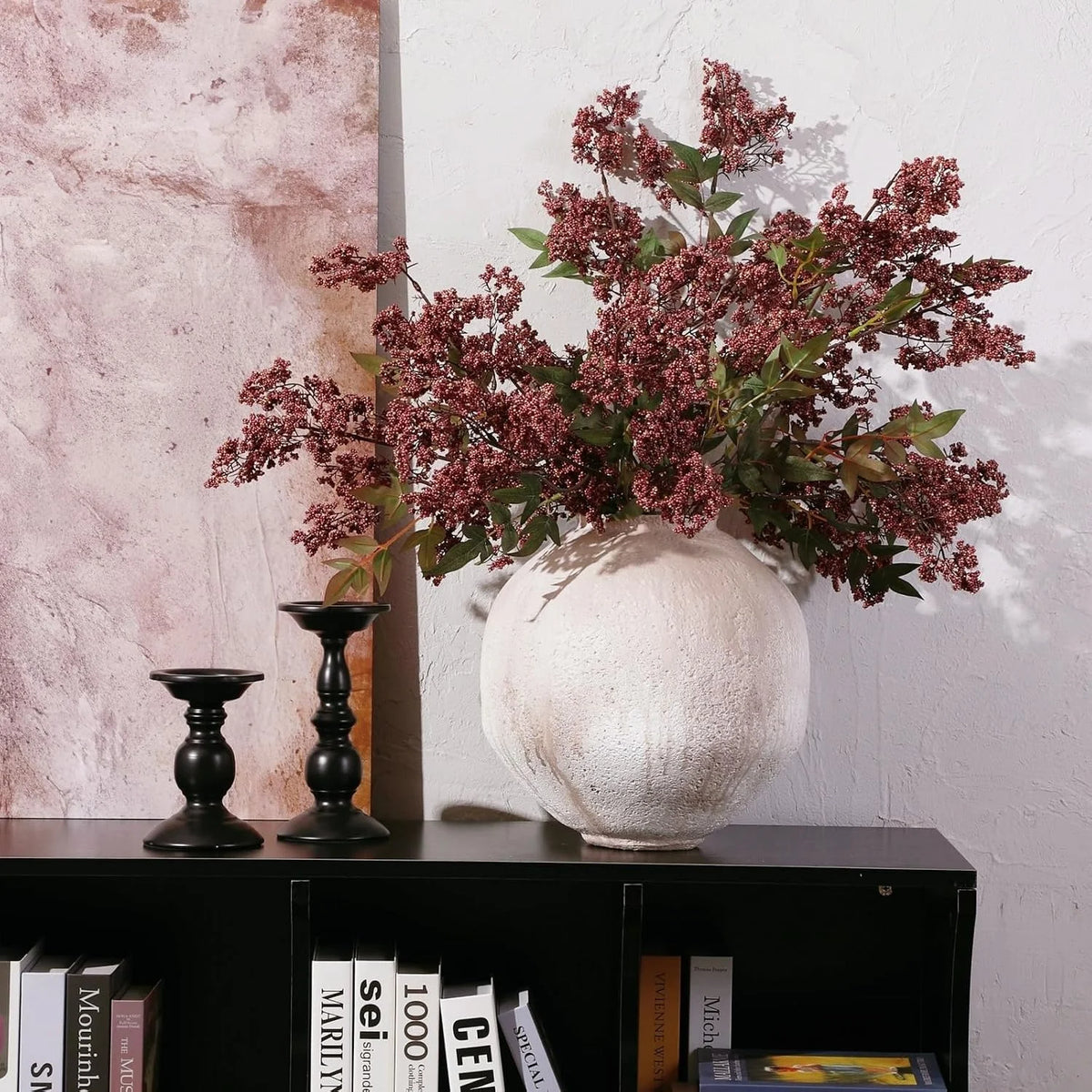 A faux branch of artificial nandina berry stem with dusty plum foliage displayed in a white vase against a textured wall.