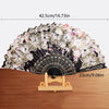 A black silk flower folding fan with pink and white floral patterns displayed on a wooden surface.